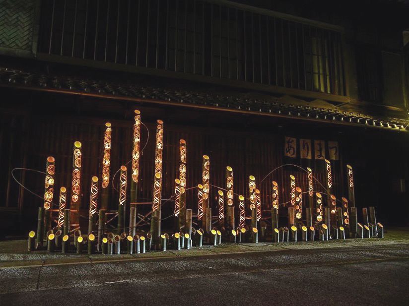夜の町家の前に、絞りの模様を彫り抜いた竹あかりが何十本も立ち並び、内側からオレンジ色の光がこぼれている