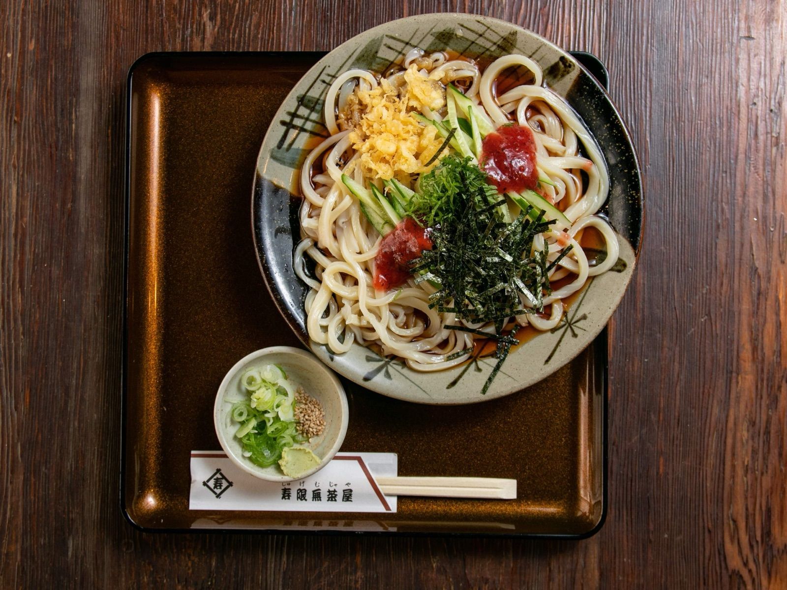 Chilled bukkake udon on a black tray, topped with tempura crumbs, cucumber, shiso, shredded nori and pickled plum